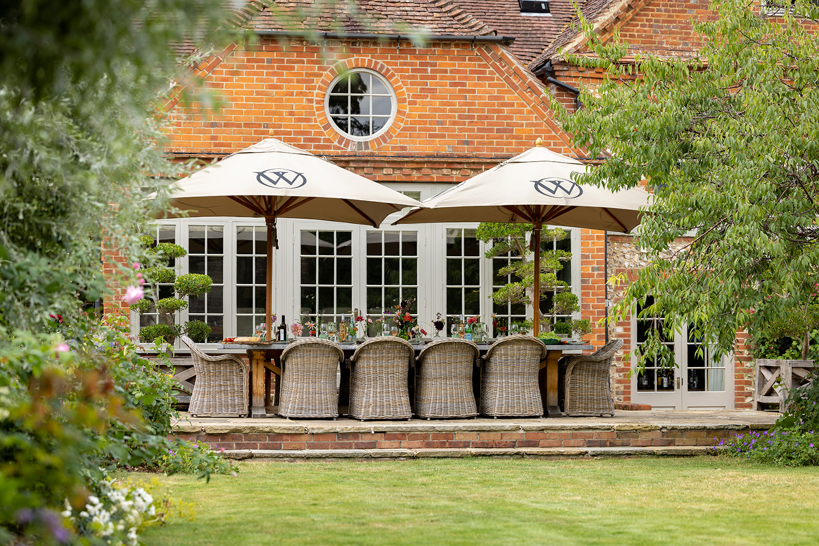 A decorated and laid outdoor dining table surrounded by woven chairs on a patio area in front of a house