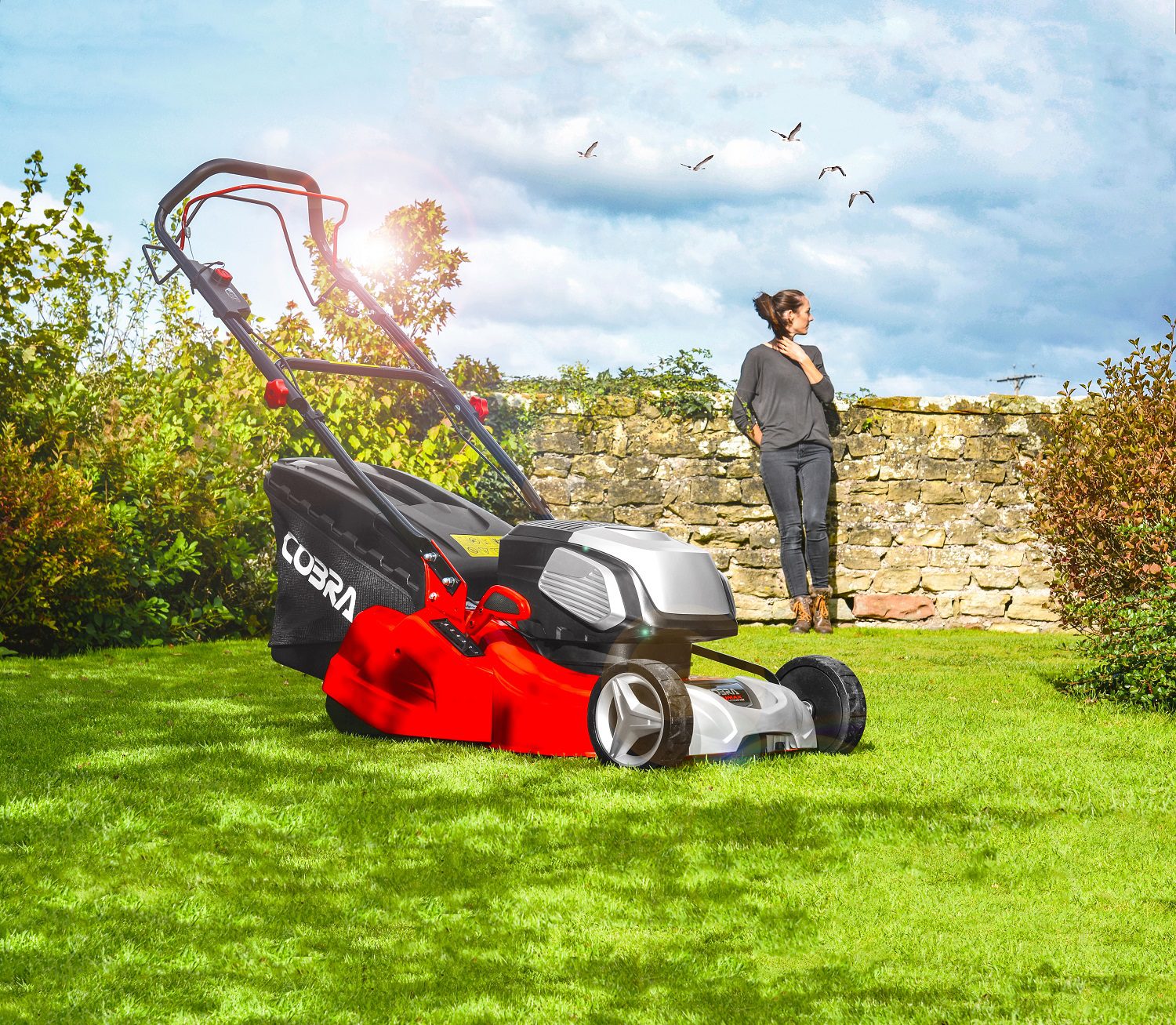 A red lawn mower on a grassy lawn, with a woman standing against a low brick wall in the background