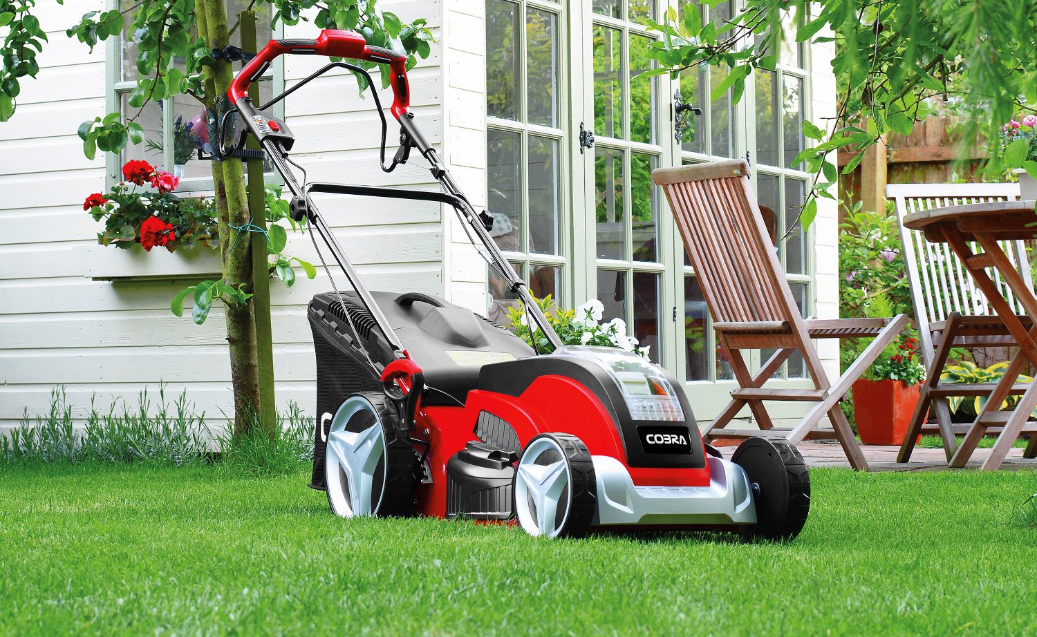 A red and black garden mower on a grassy lawn with summer seating in the background