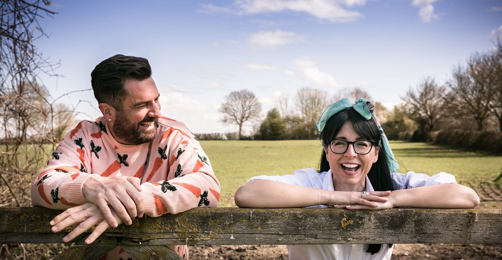 A man and woman leaning on a wooden gate smiling and laughing, with a green landscape in the background