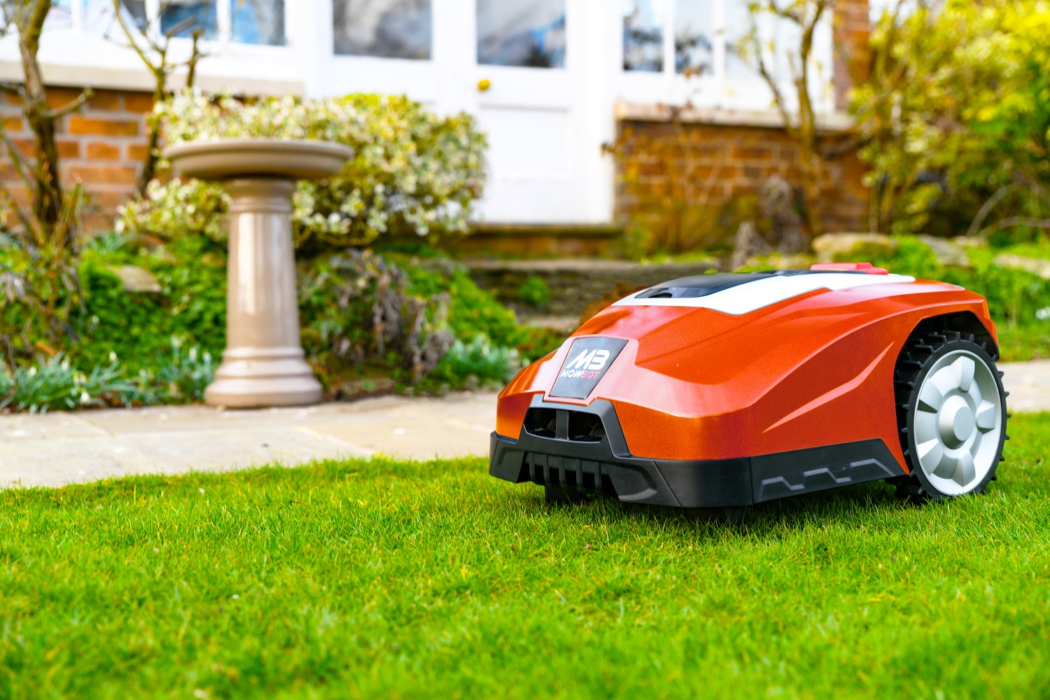 A black and red robot lawnmower on a grassy garden, with a bird bath and path in the background