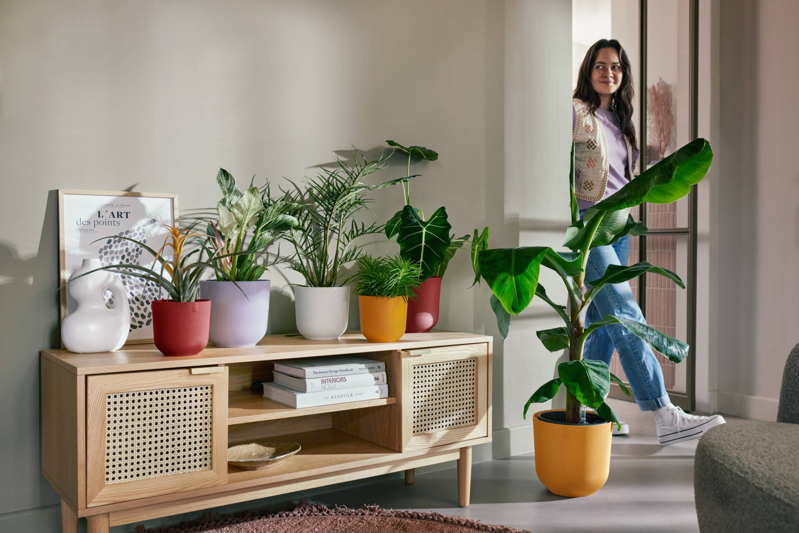a woman walking into a lounge with colourful Elho plant pots on a wooden unit