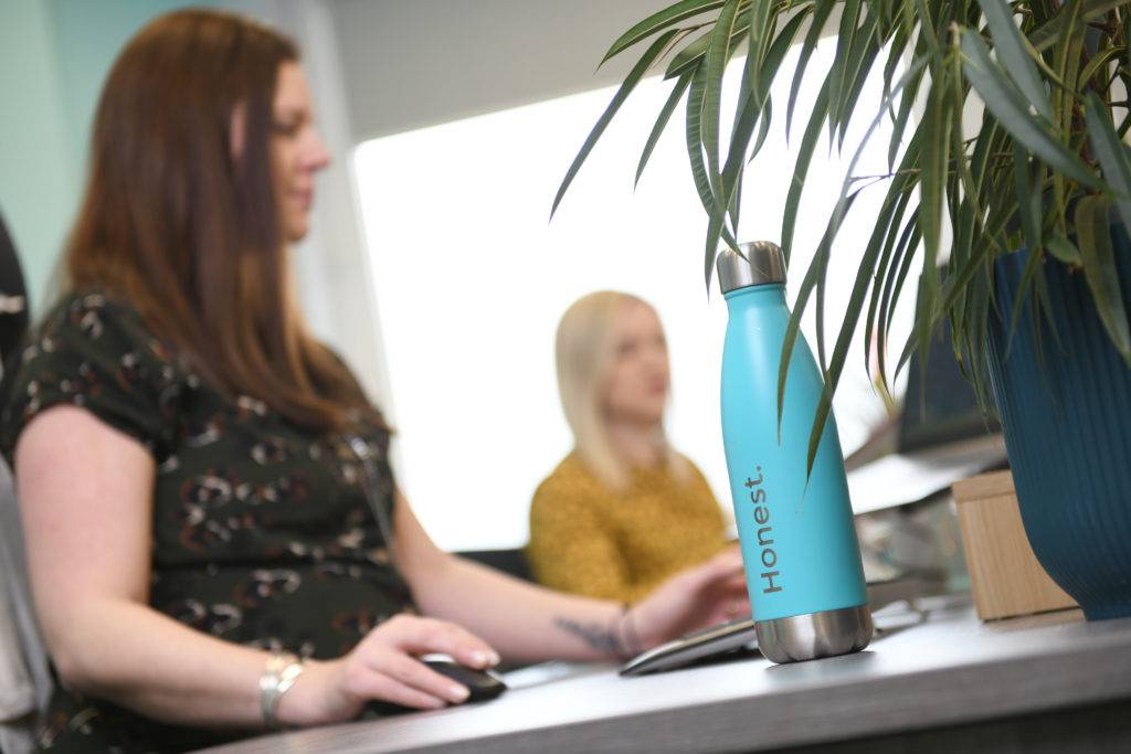 Two women working at a desk with an Honest Communications water bottle and plant in the foreground.