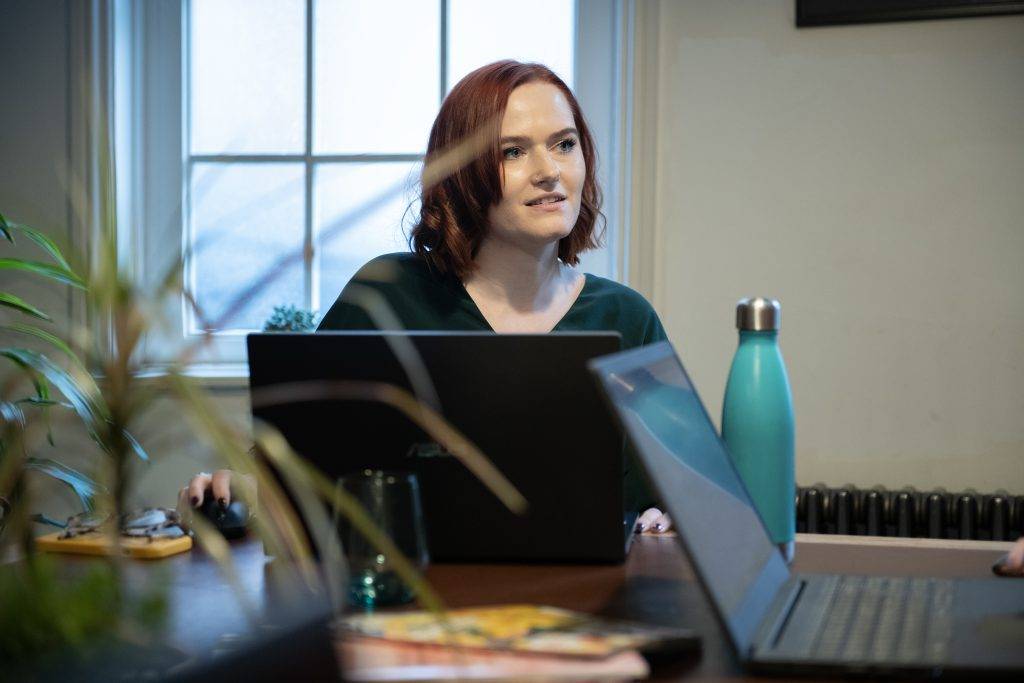 A female working at a desk with two laptops and a plant