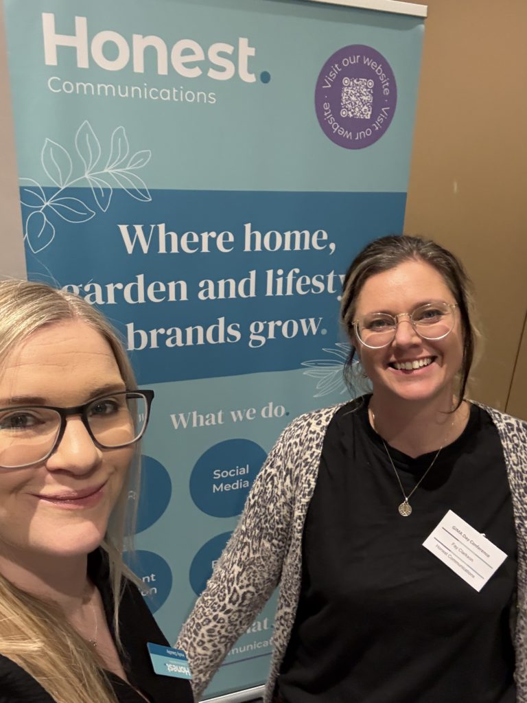 Two females smiling in front of a pull up banner for the garden PR agency Honest Communications